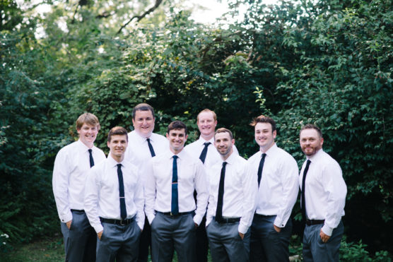 Eight men wearing white shirts and black ties stand together outdoors in front of green bushes, smiling at the camera. by Justin Salem Meyer