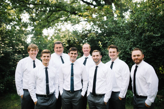 Eight men wearing white shirts and black ties stand together outdoors, smiling at the camera. They are surrounded by greenery and trees, with sunlight filtering through the leaves in the background. by Justin Salem Meyer