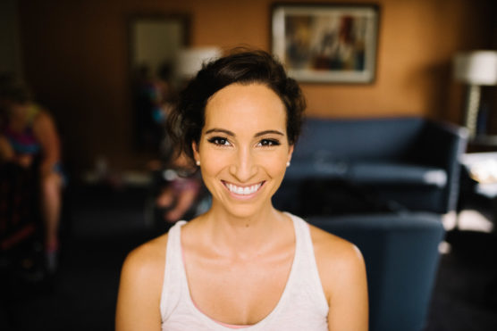 A woman with dark hair and flawless makeup smiles brightly at the camera, sitting in a warmly lit living room with blurred furniture and people in the background. by Justin Salem Meyer