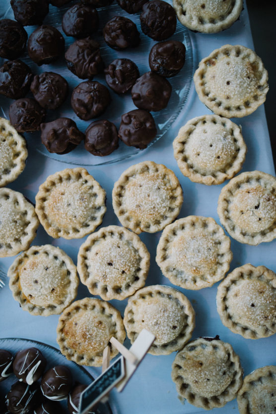 A table displays rows of small, round pies with golden, sugar-dusted crusts beside plates of chocolate-covered treats arranged neatly on clear glass dishes. by Justin Salem Meyer