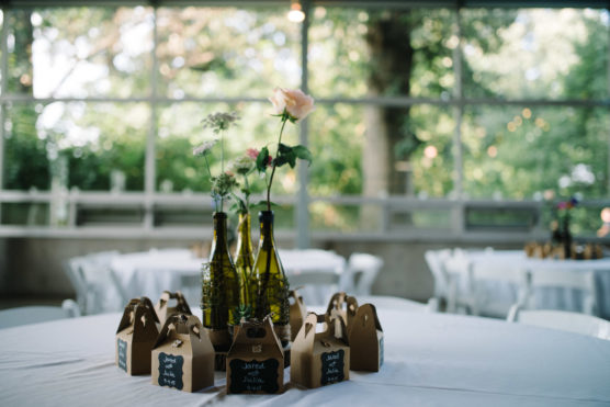 A round table set for an event, decorated with three glass bottles holding flowers at the center, and surrounded by small brown gift boxes with name tags. The background features large windows and greenery outside. by Justin Salem Meyer