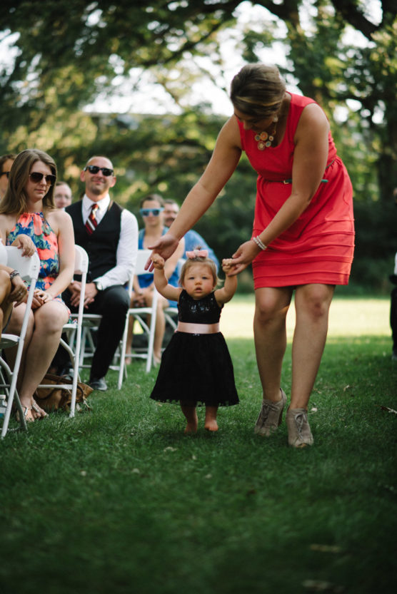 A woman in a coral dress helps a young toddler in a black dress walk down an outdoor aisle, while seated guests watch and smile. Trees and sunlight create a warm, festive atmosphere. by Justin Salem Meyer