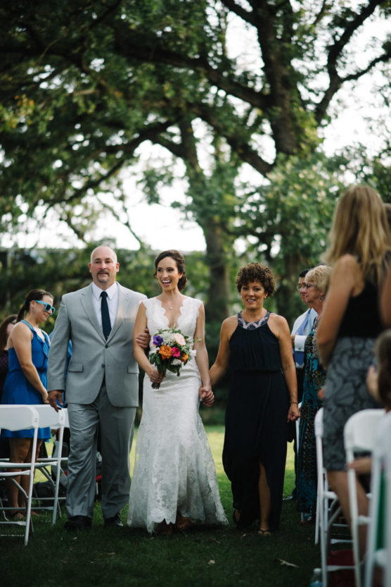 A bride in a white dress holding a bouquet walks down an outdoor aisle, smiling and holding hands with a man in a gray suit and a woman in a black dress, surrounded by seated guests and trees. by Justin Salem Meyer