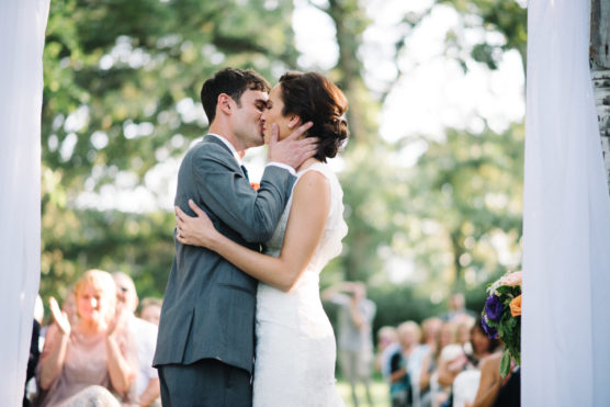 A bride and groom kiss during an outdoor wedding ceremony, surrounded by guests seated in the background under trees. The groom wears a gray suit and the bride is in a white lace dress. by Justin Salem Meyer