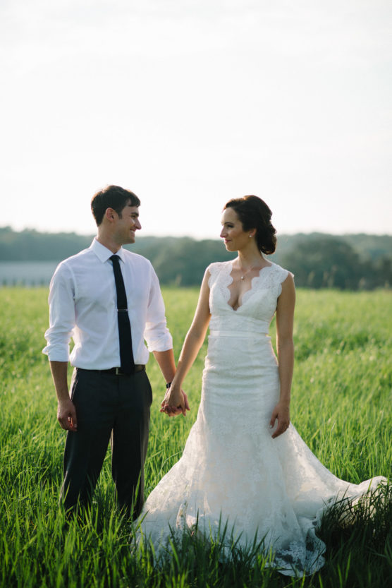 A bride in a white lace gown and a groom in a white shirt and dark tie hold hands, standing together in a lush green field with trees and a soft sky in the background. by Justin Salem Meyer