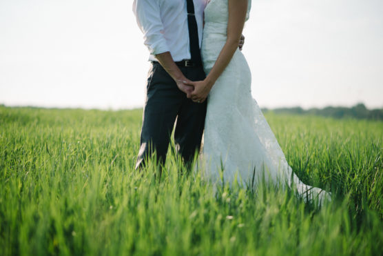A bride and groom stand in a green field, holding hands. Only their bodies from the shoulders down are visible; the bride wears a white lace dress and the groom wears dark pants, a white shirt, and a tie. by Justin Salem Meyer