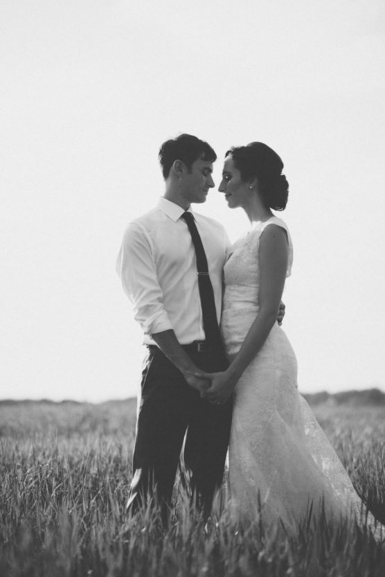 A couple dressed in wedding attire stands closely together in a grassy field, holding hands and gazing at each other, captured in black and white. by Justin Salem Meyer