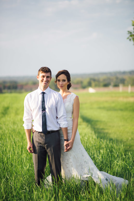 A bride in a white lace gown and a groom in a white shirt and tie stand hand in hand on a green grassy field, smiling as they pose for a wedding photo with a scenic landscape in the background. by Justin Salem Meyer