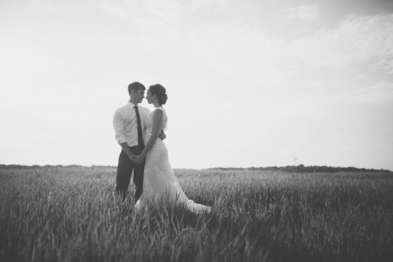 A couple in wedding attire stands holding hands in a grassy field, facing each other. The scene is in black and white with soft, natural light and a serene, open sky in the background. by Justin Salem Meyer