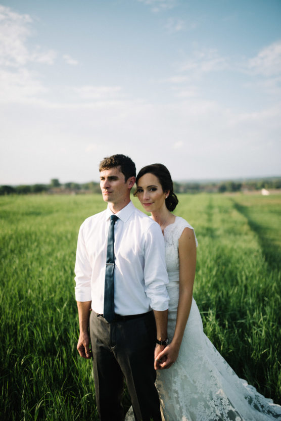 A bride in a white gown stands closely behind a groom in a white shirt and tie, holding hands in a green field under a blue sky, both looking calmly into the distance. by Justin Salem Meyer
