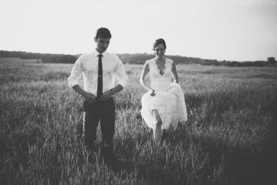 A black and white photo of a bride and groom walking through a grassy field. The bride is holding up her lace dress and smiling, while the groom adjusts his shirt and tie. The background shows open landscape and a distant treeline. by Justin Salem Meyer