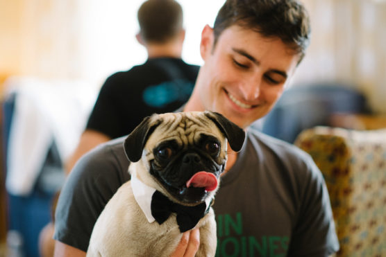 A smiling man holds a pug wearing a black bow tie. The pug is looking at the camera with its tongue out. The background is softly blurred. by Justin Salem Meyer