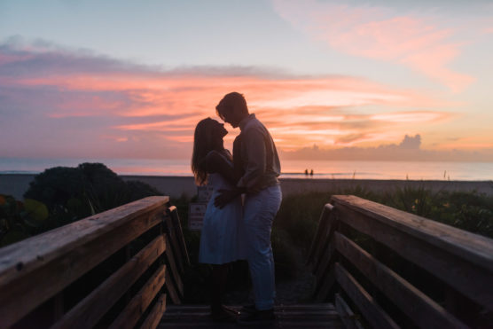 A couple stands close together on a wooden walkway, silhouetted against a vibrant sunset sky at the beach. The sky glows with pink, orange, and purple hues as they share a romantic moment. by Justin Salem Meyer