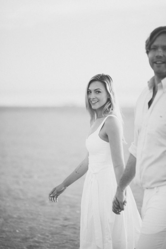 A woman in a white dress smiles and holds hands with a man, both walking on a sandy beach. The photo is black and white, with the man partially out of frame and the background softly blurred. by Justin Salem Meyer