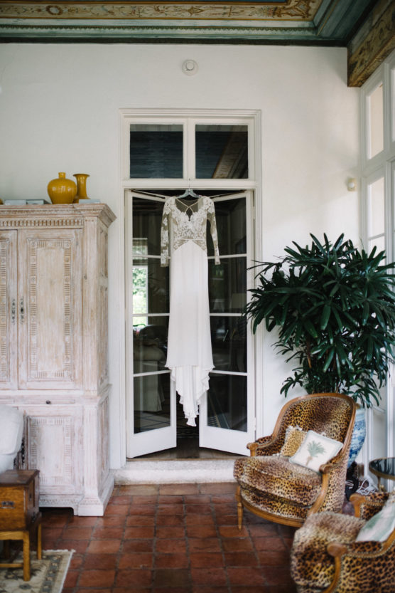 A long-sleeved, white wedding dress with lace detailing hangs on a door in a cozy room with terracotta floor tiles, a large green plant, an ornate wooden cabinet, and a leopard-print armchair. by Justin Salem Meyer