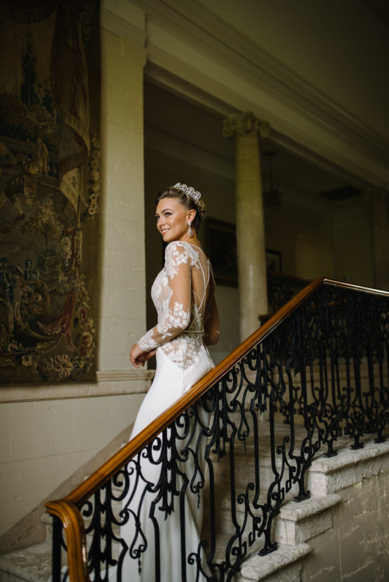 A bride in a long-sleeved lace wedding dress and jeweled headpiece smiles while walking up a grand staircase with ornate black railings inside an elegant building. by Justin Salem Meyer