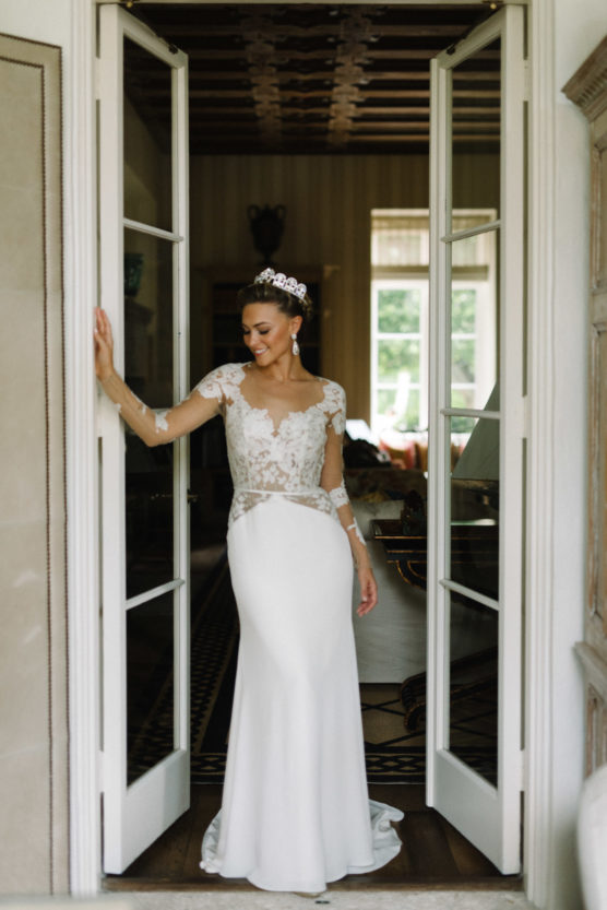 A bride in a white gown with lace detailing stands smiling in a doorway. She wears a tiara and earrings, her hair is styled up, and natural light streams in from windows behind her. by Justin Salem Meyer