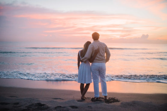 A couple stands on a sandy beach, facing the ocean and colorful sunset sky. The woman in a white dress leans on the man, who is wearing light-colored clothes. Waves gently reach the shore. by Justin Salem Meyer