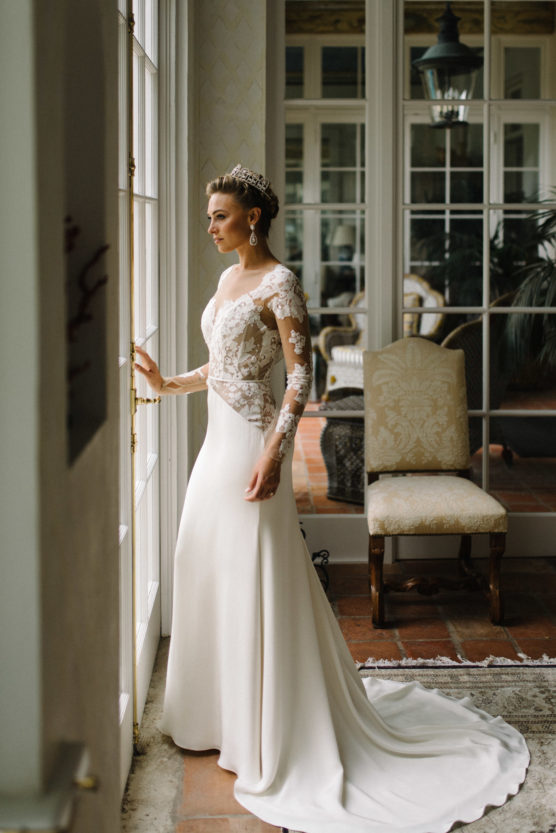 A woman in a long-sleeved, lace and satin wedding dress and tiara stands by tall windows, looking outside. She is in an elegant room with large mirrors, classic chairs, and natural light streaming in. by Justin Salem Meyer