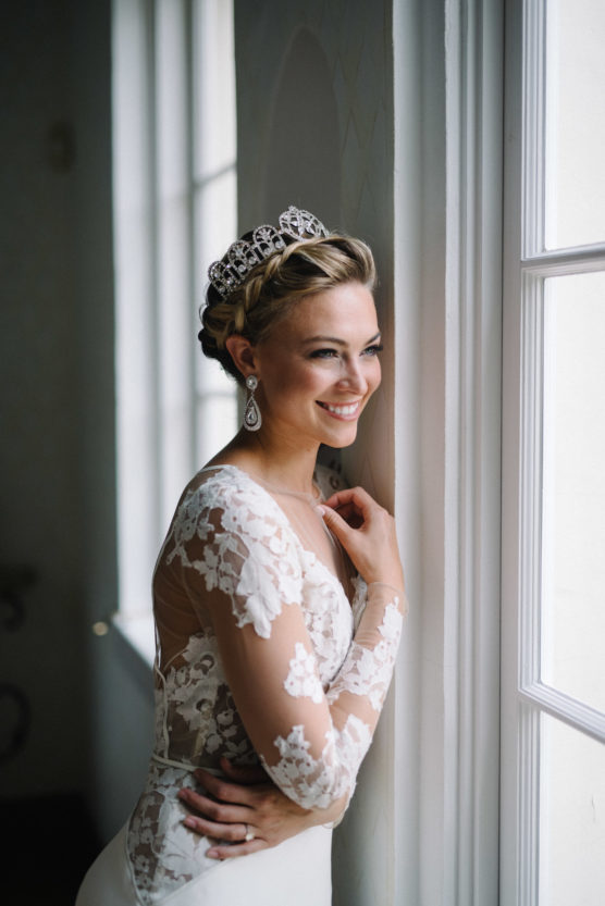 A smiling bride in a lace wedding dress and tiara stands by a window, gazing outside with her hand resting on the wall. Soft natural light highlights her elegant earrings and braided hair. by Justin Salem Meyer