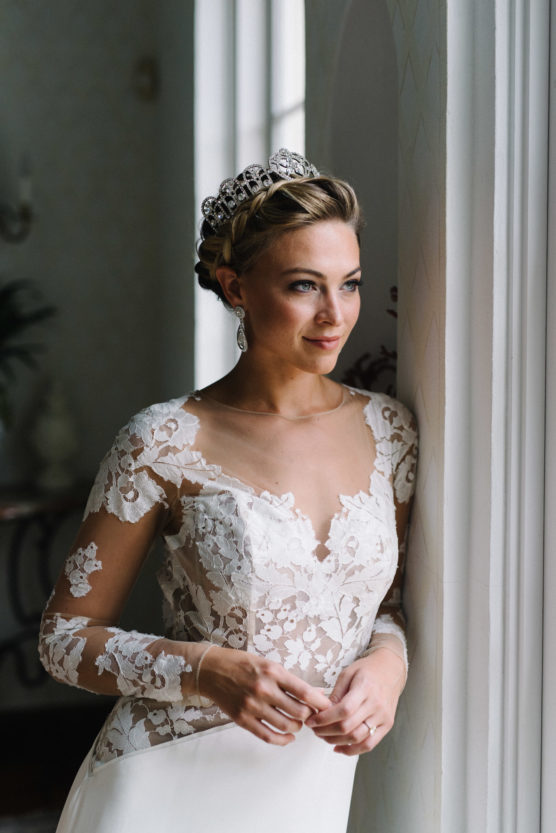A bride wearing an elegant lace wedding dress and a tiara stands by a window, looking outside thoughtfully with soft natural light illuminating her face. by Justin Salem Meyer