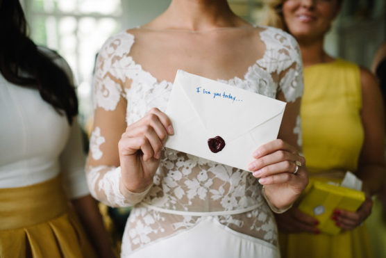 A woman in a white lace dress holds an envelope sealed with red wax. The envelope reads I love you today... Two women in yellow outfits stand nearby, slightly out of focus. by Justin Salem Meyer