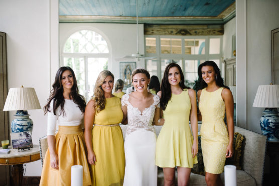 Five women stand indoors, smiling together; the woman in the center wears a white wedding dress, while the other four wear coordinating yellow outfits. The room is bright with decorative lamps, mirrors, and a blue-green ceiling. by Justin Salem Meyer