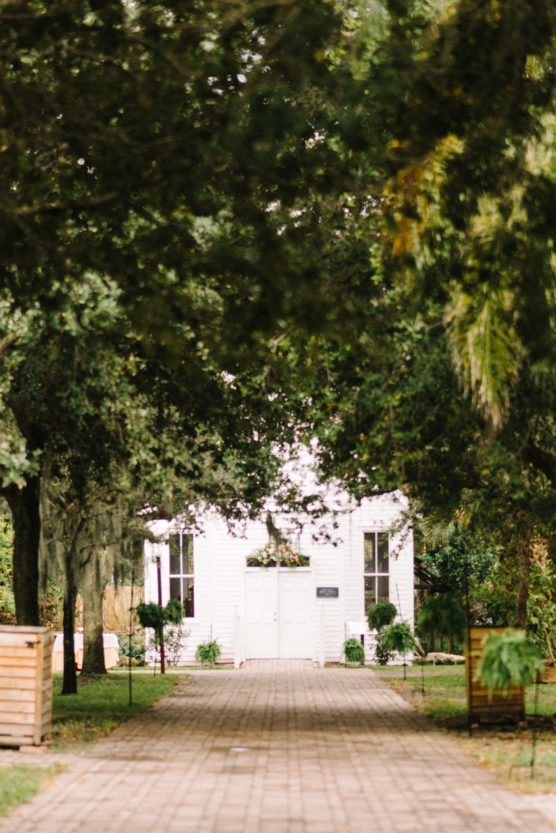 A white building with double doors sits at the end of a brick pathway, framed by tall, lush trees and greenery on both sides, creating a peaceful, inviting atmosphere. by Justin Salem Meyer
