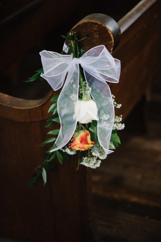 A white and an orange rose with greenery and baby’s breath are tied to a wooden pew with a sheer white ribbon, likely as a wedding or event decoration. by Justin Salem Meyer