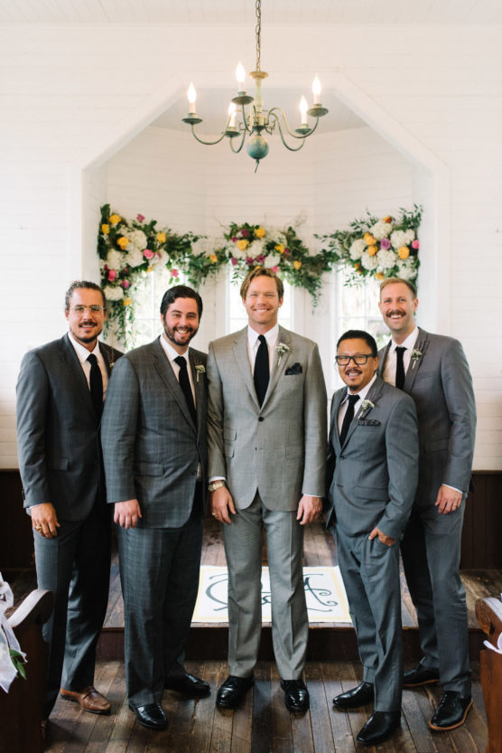 Five men in suits stand together inside a bright chapel, smiling for a photo. Floral arrangements and a chandelier decorate the space behind them, creating a festive and elegant atmosphere. by Justin Salem Meyer