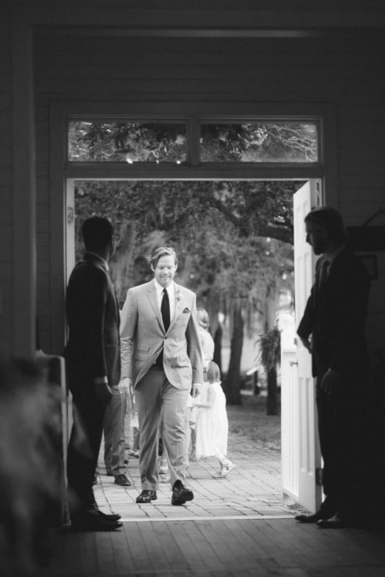 A man in a light suit and tie walks through an open doorway, smiling. Two men in dark suits stand on either side of the entrance. A woman and child are visible outside in the background. The image is black and white. by Justin Salem Meyer