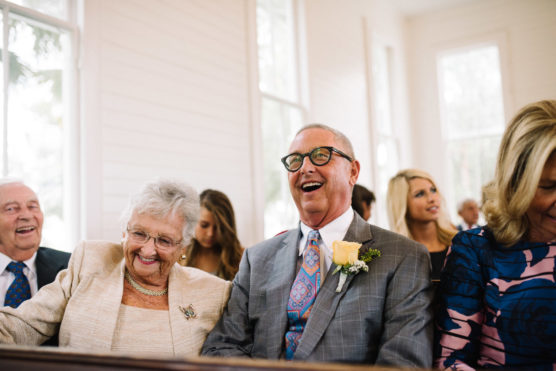 An elderly man and woman sit together at a wedding ceremony, smiling and laughing. The man wears glasses, a suit, and a yellow rose boutonniere; the woman wears a light-colored outfit. Other guests are seated in the background. by Justin Salem Meyer