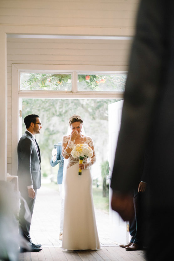 A bride in a white dress holds a bouquet and wipes a tear from her eye as she stands in a doorway. A man in a suit stands nearby, with others partially visible around them. Light streams in from outside. by Justin Salem Meyer