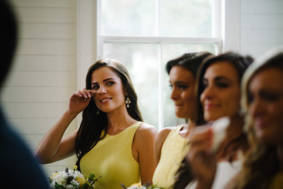 A woman in a yellow dress wipes a tear from her eye, standing beside three other women indoors near a window. All appear dressed up, possibly for a special occasion like a wedding. by Justin Salem Meyer
