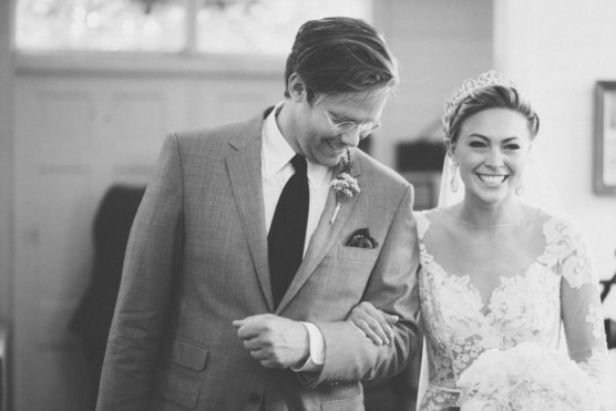 A joyful bride and a man in a suit walk arm in arm indoors. The bride is smiling widely, wearing a lace dress and tiara, while the man wears glasses and a suit with a boutonniere. The image is in black and white. by Justin Salem Meyer