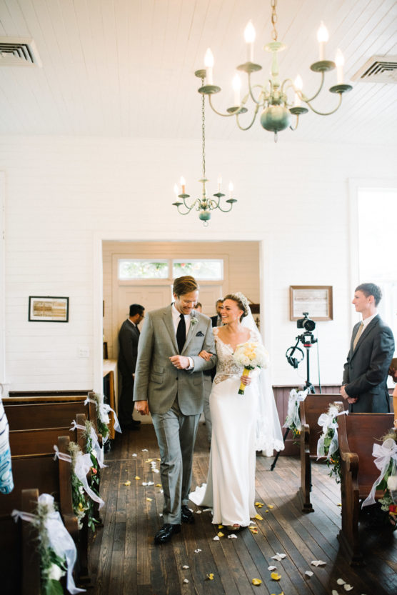 A bride in a white dress and a groom in a gray suit walk arm in arm down the aisle of a bright, decorated chapel, smiling at each other, with guests and a photographer in the background. by Justin Salem Meyer