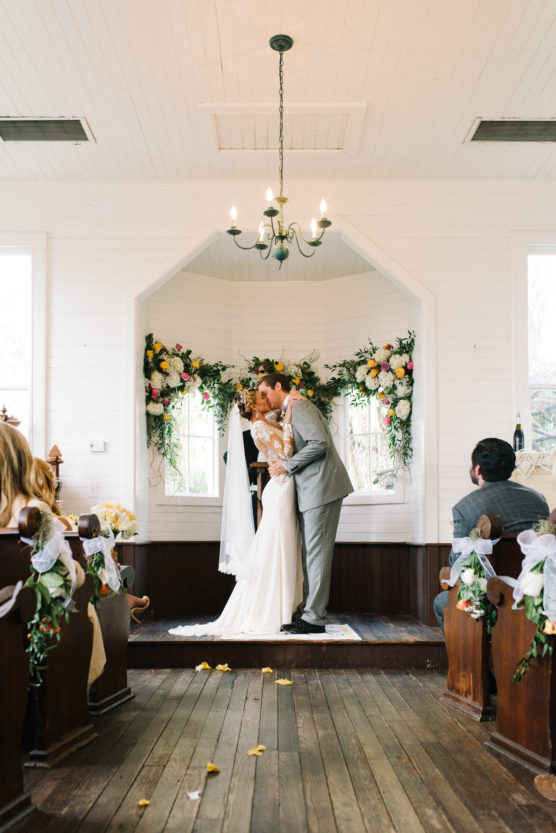 A bride and groom share their first kiss at the altar in a small, flower-decorated chapel, surrounded by guests seated in wooden pews. Light streams through windows, and flower petals are scattered on the wooden floor. by Justin Salem Meyer