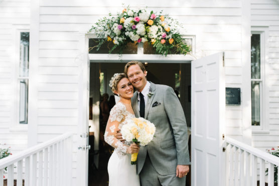 A bride in a white dress and a groom in a gray suit smile while standing in front of a white church doorway decorated with a colorful floral arrangement. by Justin Salem Meyer