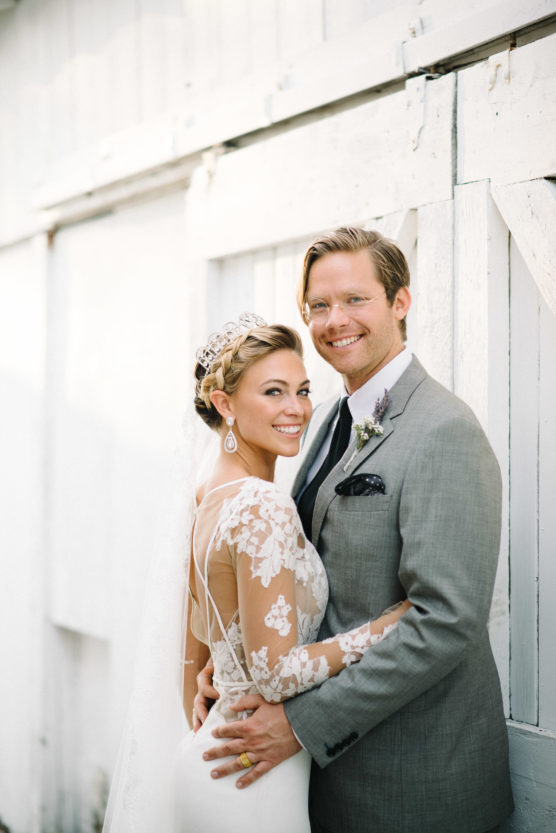 A bride in a lace wedding dress and tiara stands smiling beside a groom in a light gray suit and glasses. They are embracing in front of a white wooden barn door. by Justin Salem Meyer