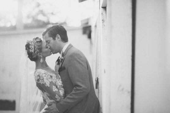 A bride and groom share a romantic kiss outdoors on their wedding day. The bride wears a lace dress, veil, and tiara, while the groom is in a suit. The photo is black and white, capturing an intimate moment. by Justin Salem Meyer