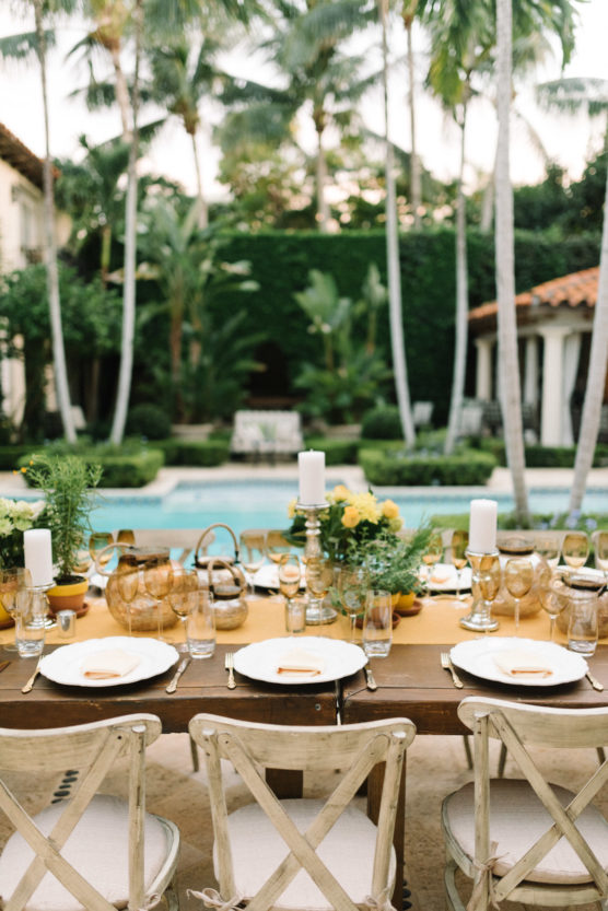 An outdoor dining table set with white plates, glasses, candles, and greenery, positioned poolside with palm trees and lush foliage in the background. The setting exudes a tropical and elegant atmosphere. by Justin Salem Meyer