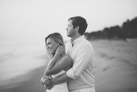 A couple stands on a beach, embracing and smiling. The image is in black and white, with the ocean and trees blurred in the background. by Justin Salem Meyer