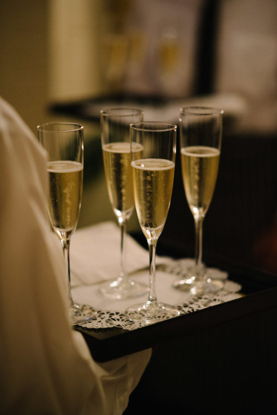 A waiter carrying a tray with five filled champagne flutes on a decorative doily, ready to be served at an event. by Justin Salem Meyer