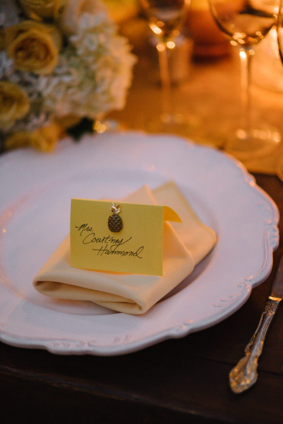 A white plate with a folded cream napkin holds a yellow place card reading Mrs. Courtney Hammond, decorated with a small gold pinecone. Glasses, cutlery, and yellow flowers are visible in the background. by Justin Salem Meyer