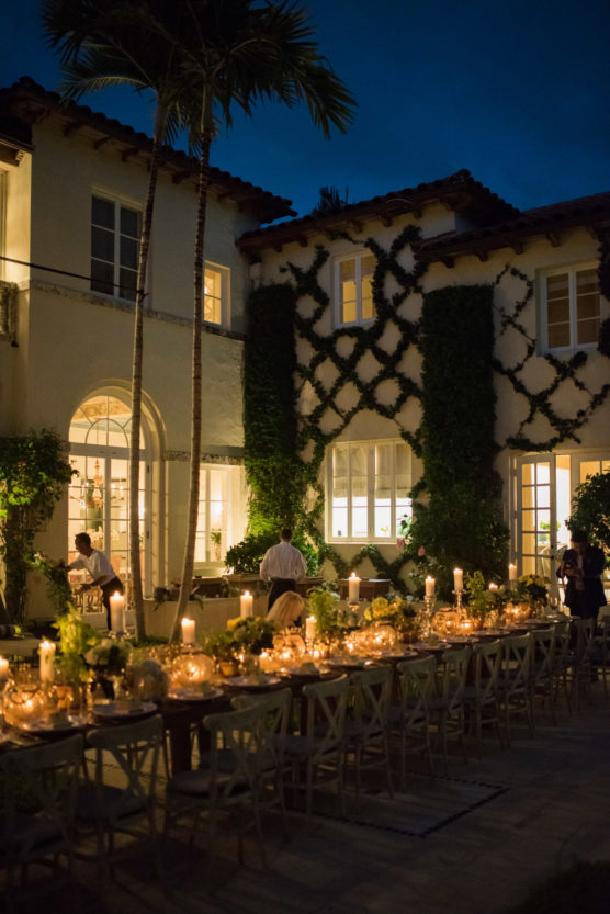A long, elegant outdoor dining table with candles and flowers is set up at night in a courtyard of a Mediterranean-style house with ivy-covered walls and tall palm trees. by Justin Salem Meyer