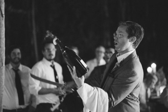 A man in a suit opens a champagne bottle with a saber, with foam spraying out, while several people in dress shirts watch in the background. The scene is lively and appears to be at a celebration or event. by Justin Salem Meyer