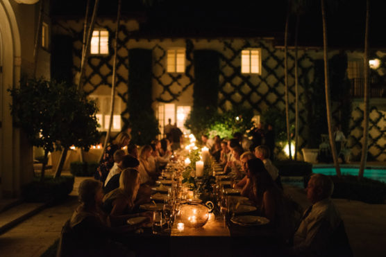A large group of people sits around a long, candlelit dinner table outdoors at night, surrounded by plants and a building covered in ivy, creating a warm, intimate atmosphere. by Justin Salem Meyer