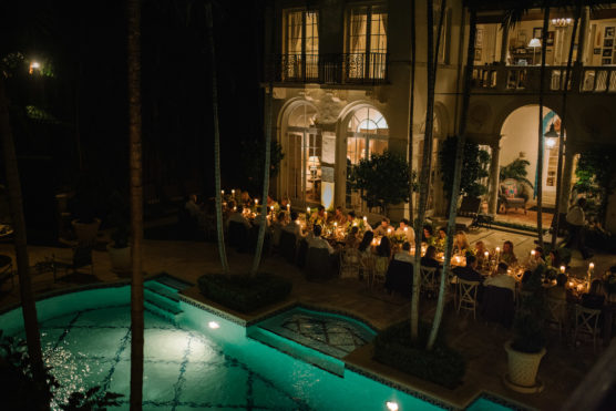 A nighttime outdoor dinner party is held beside a lit swimming pool, with long tables set for many guests and warm, glowing lights illuminating the scene in front of an elegant, multi-story house. by Justin Salem Meyer