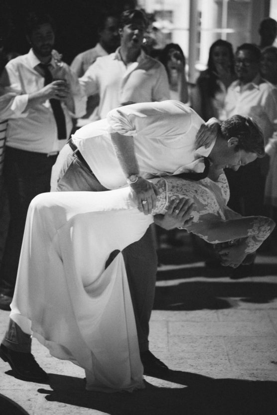 A man in a white shirt and dark pants dips a woman in a long, white dress while dancing, surrounded by a group of people watching and smiling. The scene is in black and white. by Justin Salem Meyer