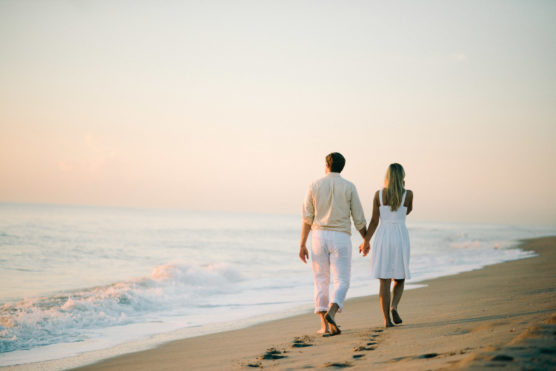A couple holding hands walks barefoot along the beach at sunset, with gentle waves rolling onto the sand and soft, warm light illuminating the scene. by Justin Salem Meyer